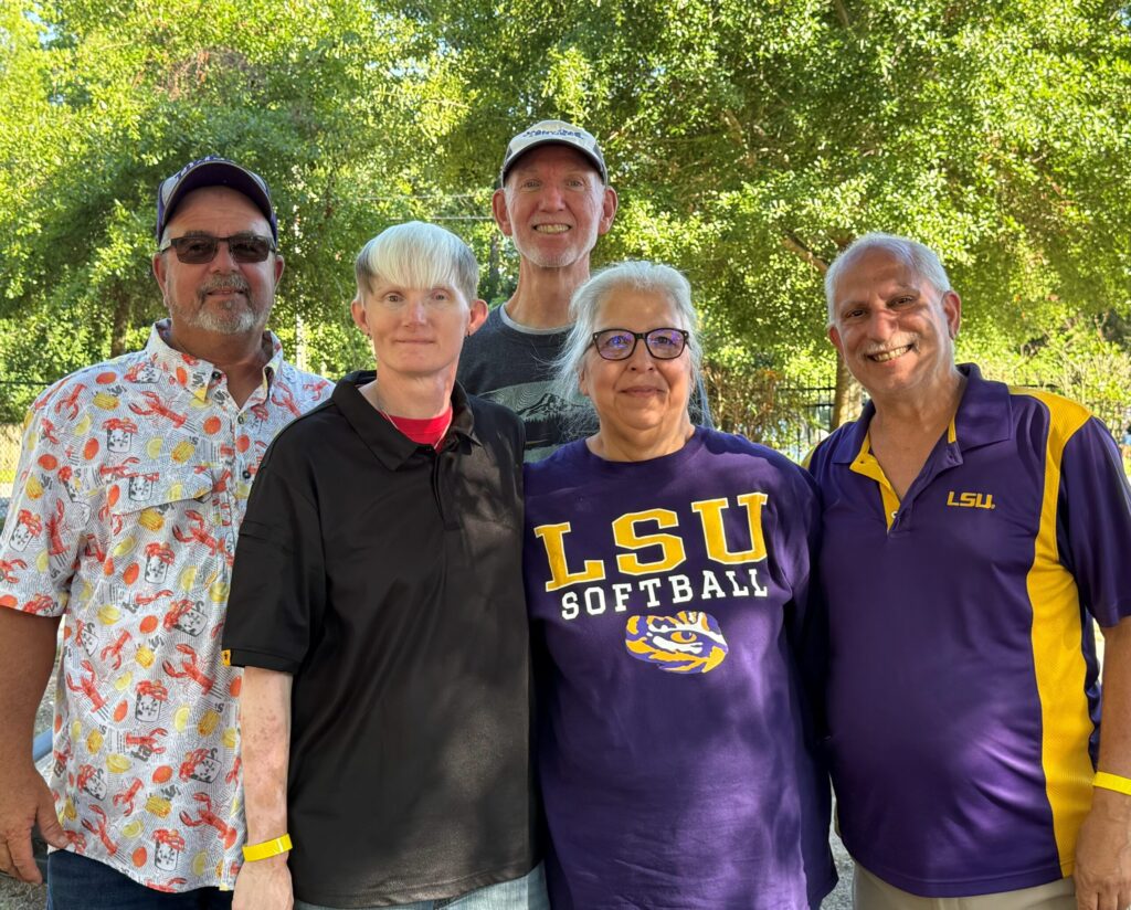 Five adults posing outdoors under leafy trees, smiling at the camera; several wear LSU softball apparel in purple and gold colors.